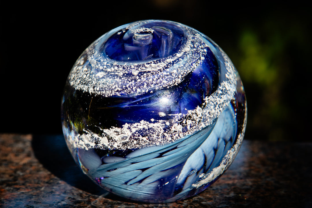 Deep blue and black swirl glass orb with cremation ash specks, sits on a stone countertop.