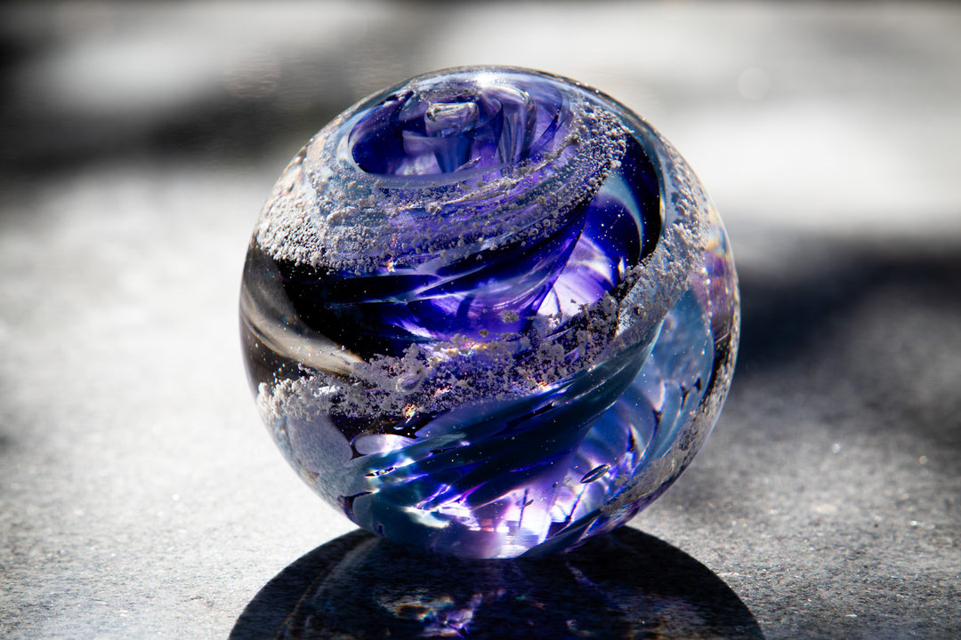 Vibrant blue and purple glass orb infused with cremation ash swirls on a sunlit surface.