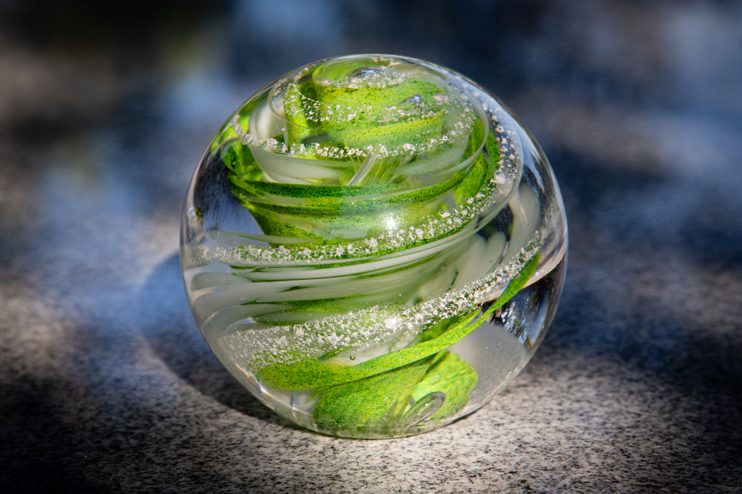Green swirled glass paperweight with ash inclusions resting on a stone surface
