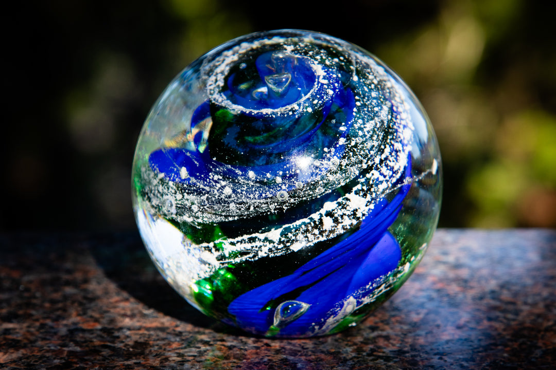 Blue and green glass sphere with cremation ash bands and white speckles outdoors on granite.