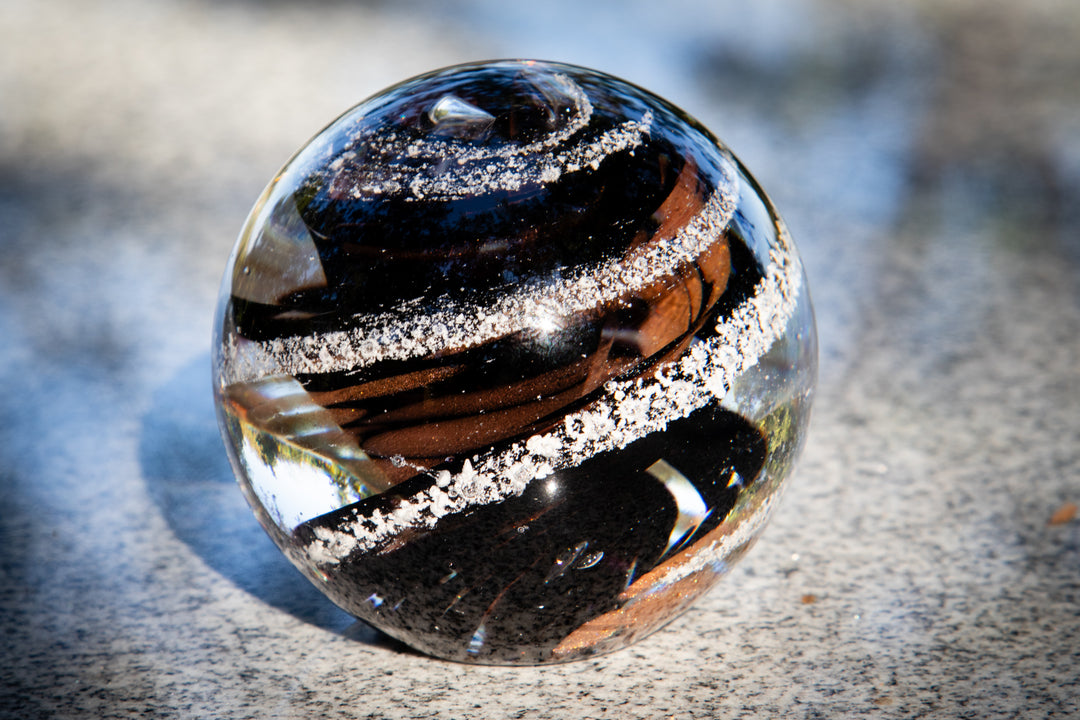 Close up of a glass tranquil swirl orb with dark brown and black swirls and white specks on a granite surface