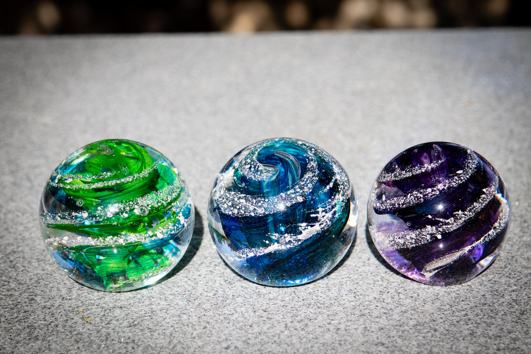 Three glass orbs in green blue and purple with white ash swirls on a gray surface.
