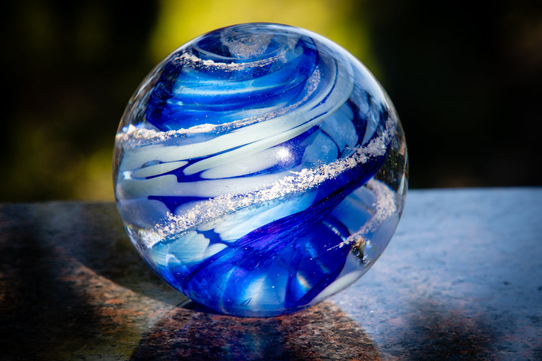 Blue glass Sparkle Orb with swirling cobalt and azure bands and white glitter accents on a granite surface
