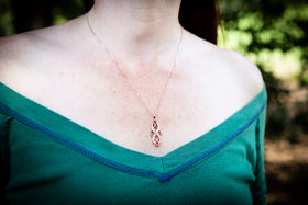 A close-up of a person wearing a delicate rose gold cremation necklace with a small pendant, paired with a green off-the-shoulder top, set against a blurred outdoor background.