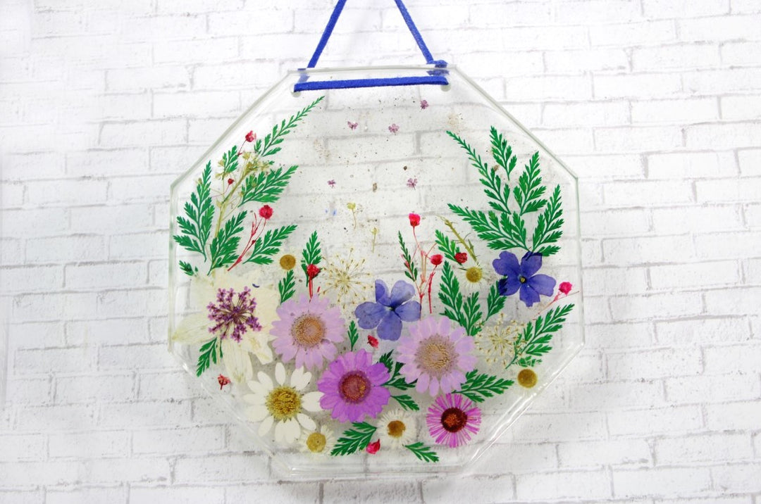 Clear octagonal glass wall ornament with a colorful pressed flower arrangement and ashes, hung by a blue ribbon.