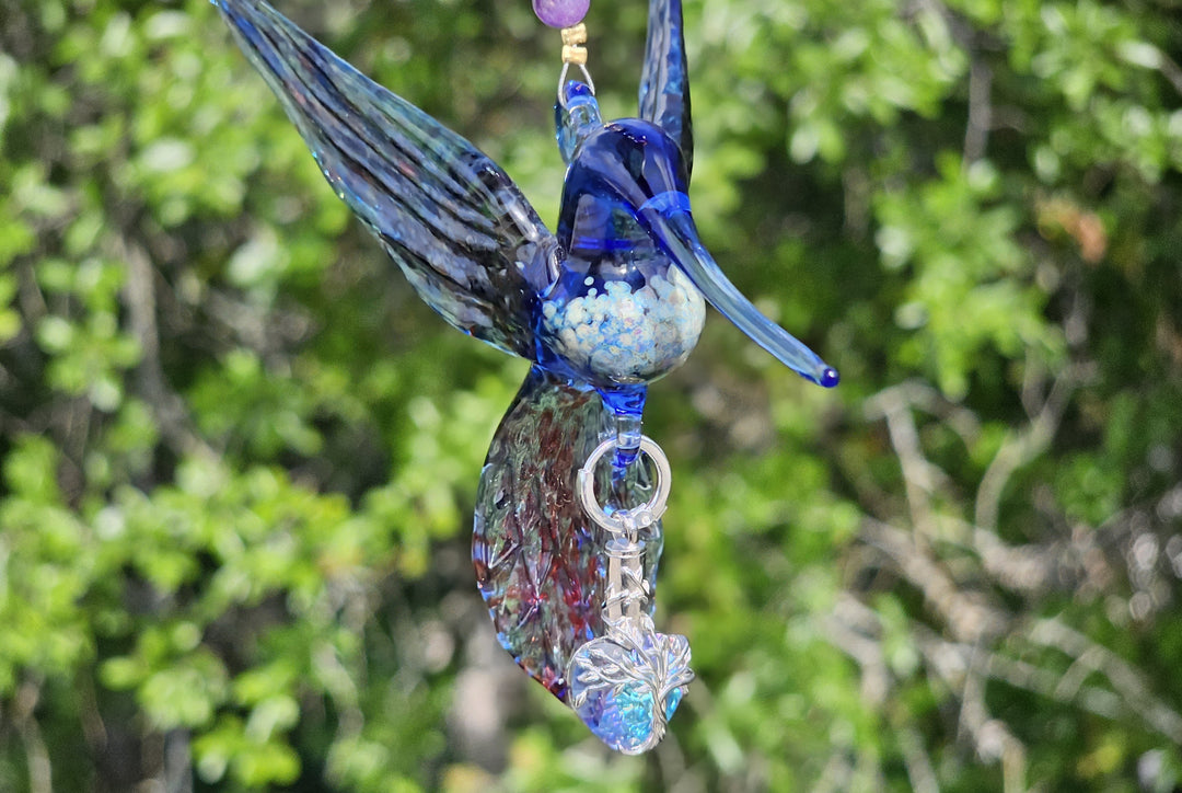 Blue glass hummingbird pendant with a crystal leaf charm on a beaded strand against green background.