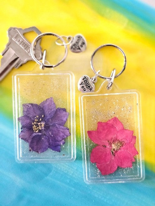 Two flower keychains in clear frames holding dried purple and pink flowers attached to metal key rings.