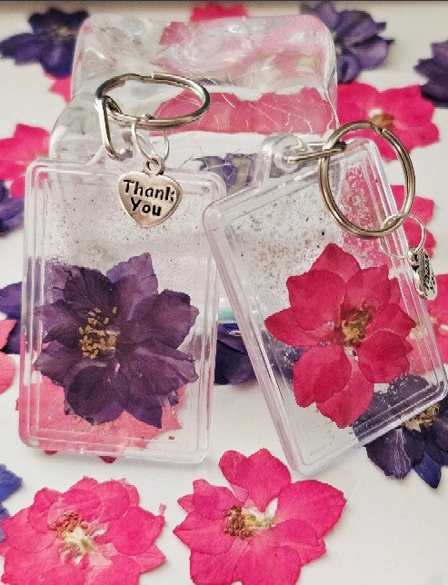 Two cremation ash flower keychains in clear frames featuring a purple and a pink dried flower, each with a Thank You charm.