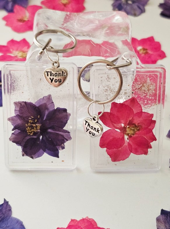 Two clear plastic flower keepsake keychains with purple and pink dried flowers and Thank You charms
