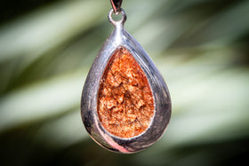 Teardrop-shaped pendant with orange center and silver frame against a blurred green background with hair from a person