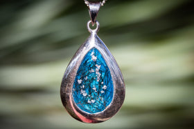 Teardrop-shaped pendant with blue center and silver frame against a blurred green background with hair from a person