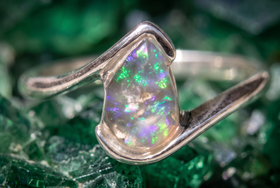 A silver cremation ring featuring a teardrop-shaped opal gemstone with vibrant green, blue, and purple flashes, set against a textured green crystal background.