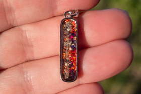 A close-up of a hand holding a small rectangular pendant with cremation ash with a silver-toned frame and a colorful, glittery orange, red, and purple inlay.
