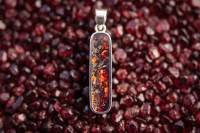 A silver-toned vertical cremation pendant necklace featuring a rectangular inlay with fiery orange, red, and black marbled patterns, set against a blurred background of dark red gemstones.