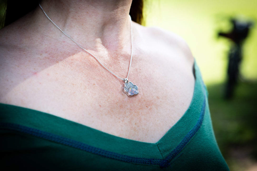 A close-up lifestyle shot of a woman wearing a silver necklace with a small, textured pawprint shaped cremation pendant. She is wearing a dark green off-the-shoulder top, with a blurred outdoor background.