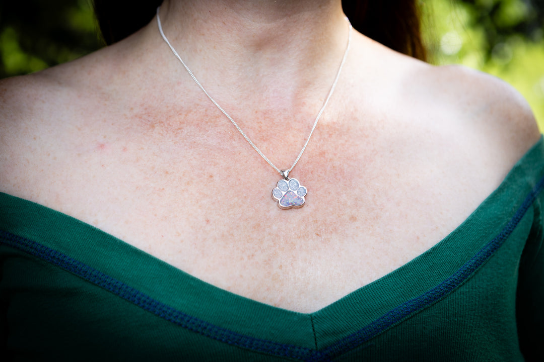 A close-up of a person's neck and chest wearing a silver paw-shaped cremation pendant necklace on a thin chain, paired with a dark green off-the-shoulder top.