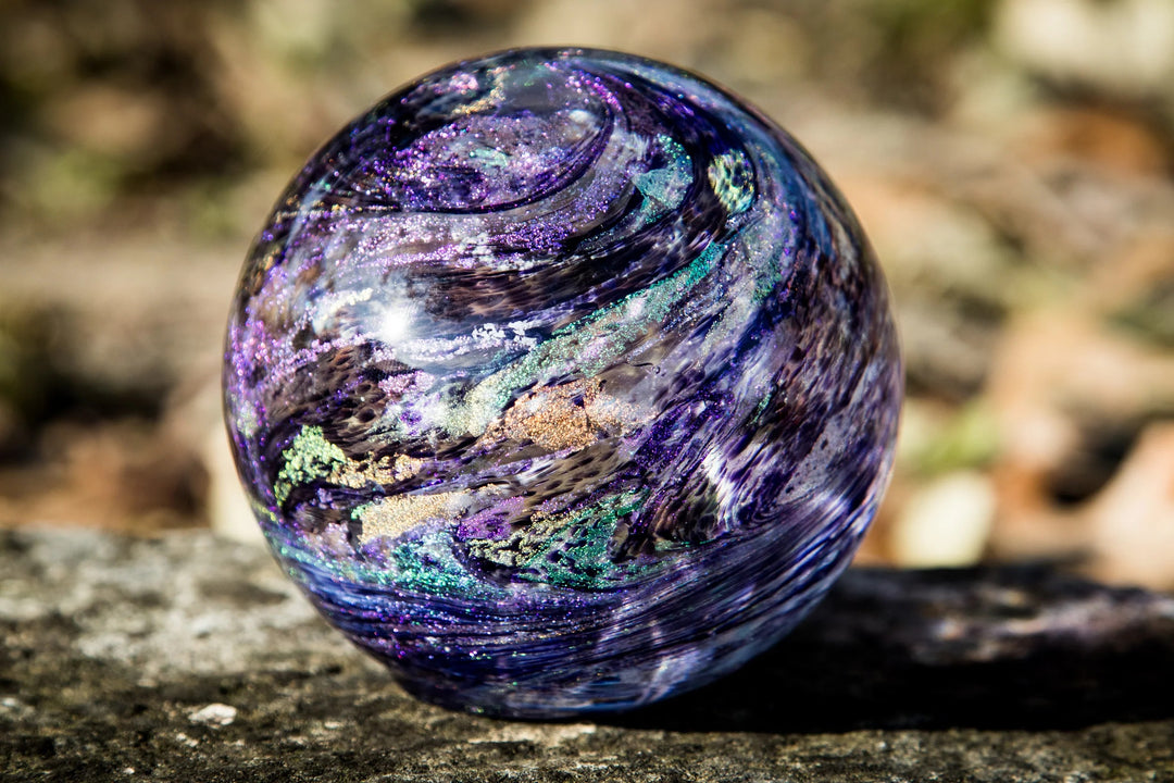 A polished iridescent glass cremation sphere with swirling purple, blue, green, and gold patterns, resting on a stone surface with a blurred natural background.
