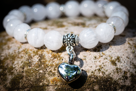 A beaded bracelet with white marbled gemstone beads, silver spacer rings, a silver floral charm, and a silver heart-shaped pendant with a green-blue iridescent center, displayed on a textured stone surface.
