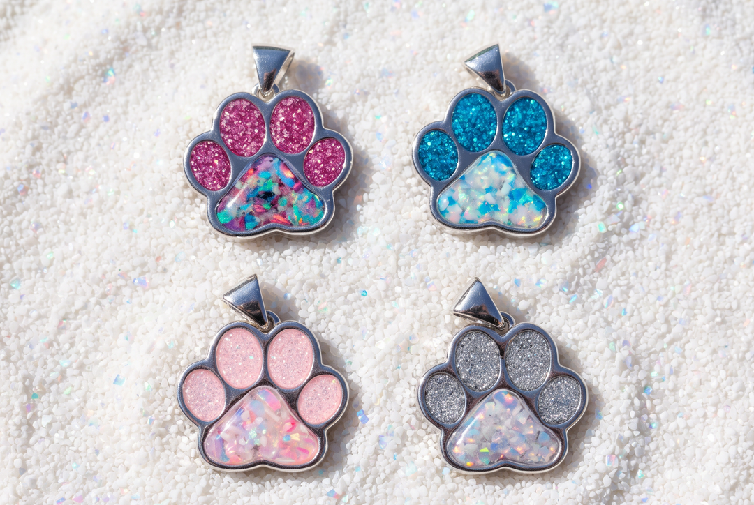 Four paw-shaped pawprint cremation pendant charms with glitter and opal-like inlay details, arranged in a 2x2 grid on a textured white surface. The paws feature pink, blue, light pink, and silver glitter designs.