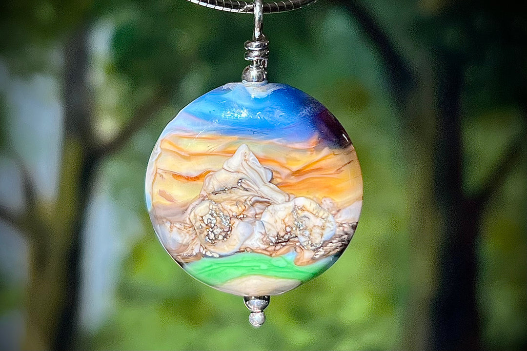Round glass pendant depicting a mountain sunset with blue sky and orange hills cremains encased inside