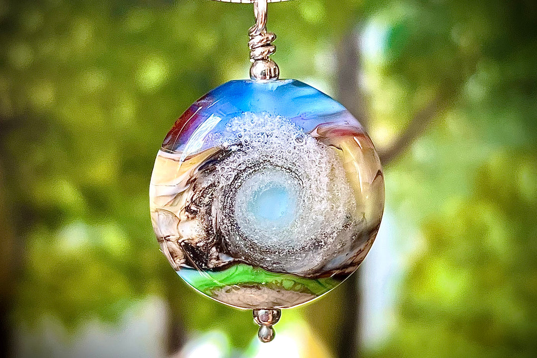 Round glass pendant with a pastel mountain sunset and iridescent glow, cremation ash visible on both sides.