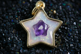 A star-shaped pendant charm with a gold-tone metal border, featuring an iridescent white opal inlay and a small purple amethyst star in the center, resting on a bed of dark blue and black iridescent pebbles.