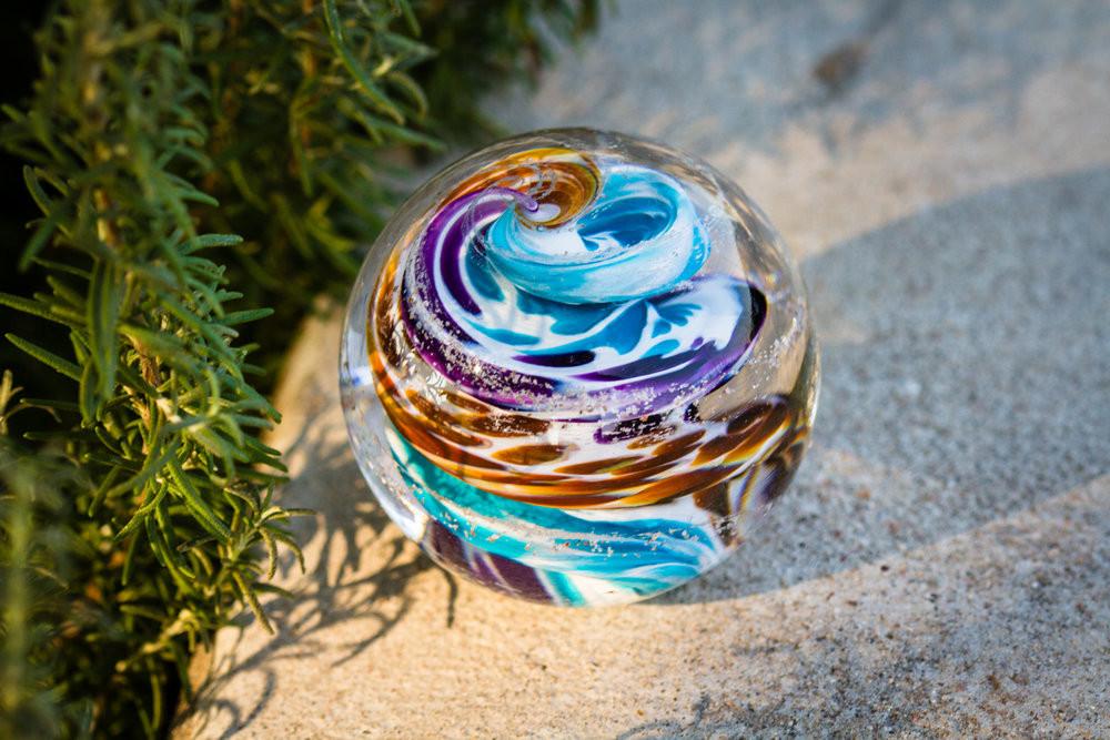 Overhead view of a multicolor glass cremation orb with cremains, blue white yellow and purple swirls on sandy ground