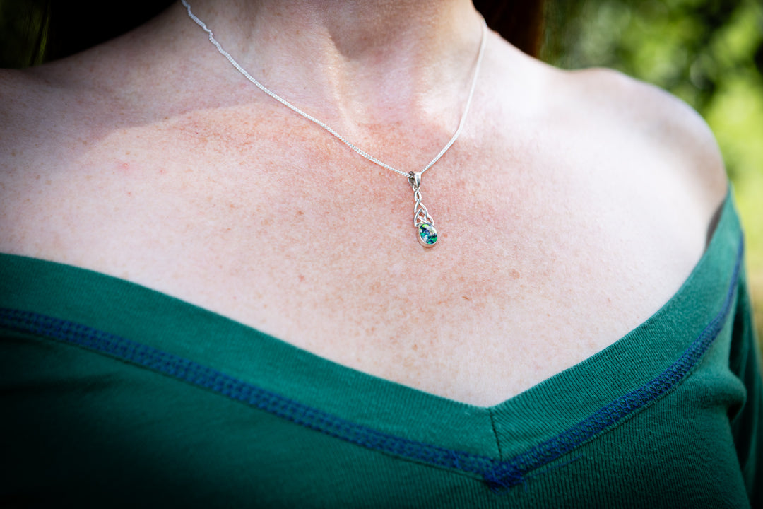 A close-up of a person's neck and upper chest wearing a delicate silver chain cremation necklace with a small teardrop pendant featuring a green stone, paired with a dark green off-the-shoulder top.