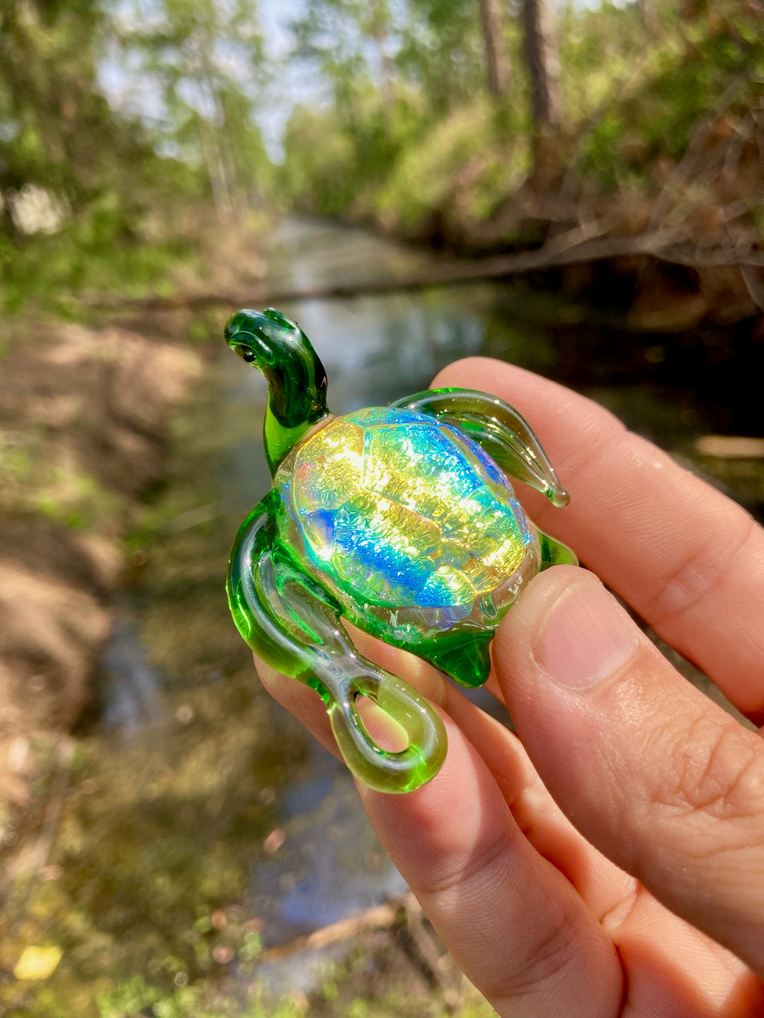 Dichroic glass sea turtle pendant with iridescent shell held between fingers outdoors beside a stream.