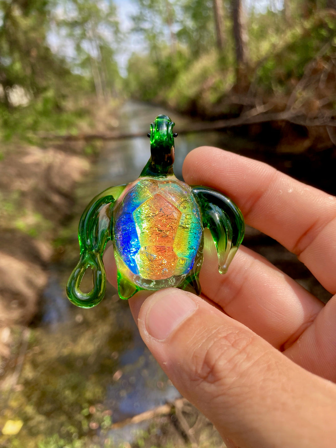 Dichroic glass sea turtle pendant in green with iridescent shell shown from the side, held outdoors by a stream.