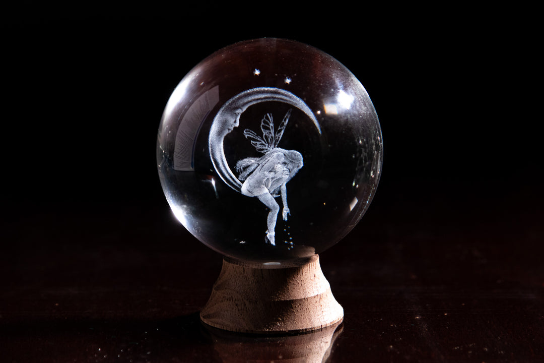 Clear crystal marble showing a fairy on a crescent moon among stars on a wooden stand.