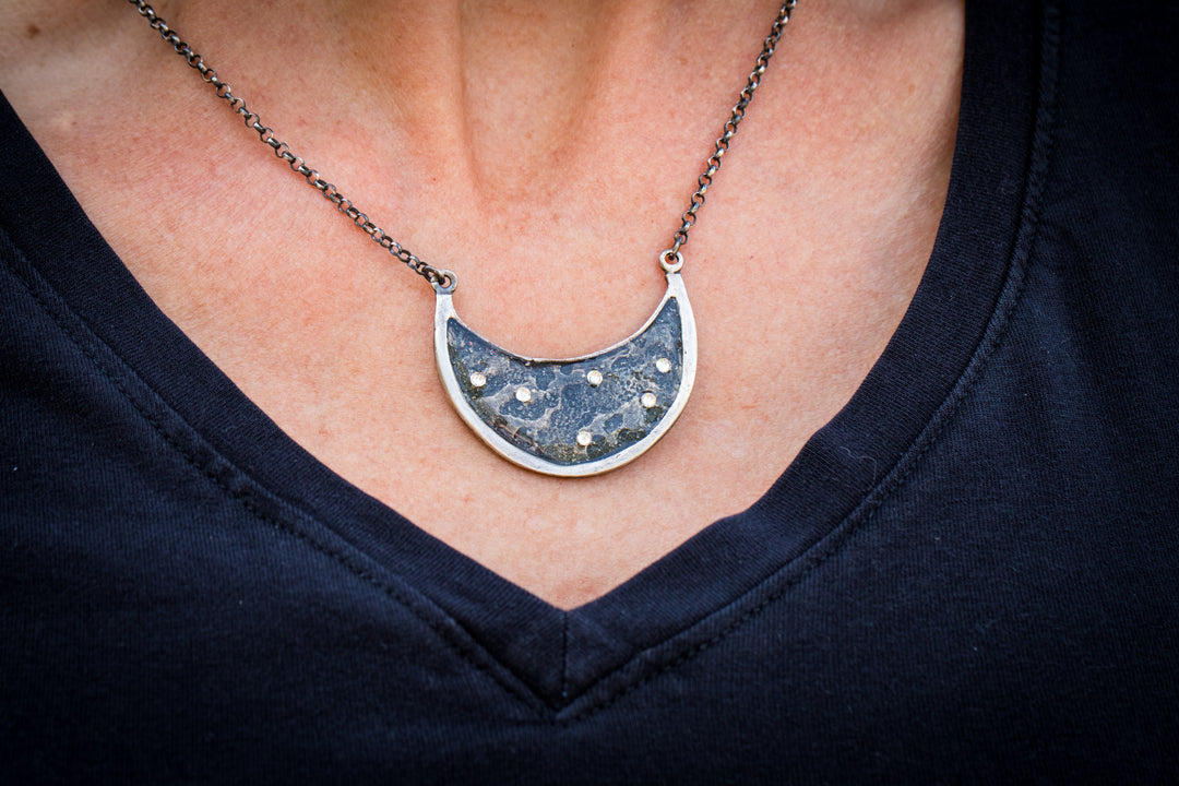Silver crescent moon pendant with blue resin inlay and scattered crystal stars on a chain against a dark top.