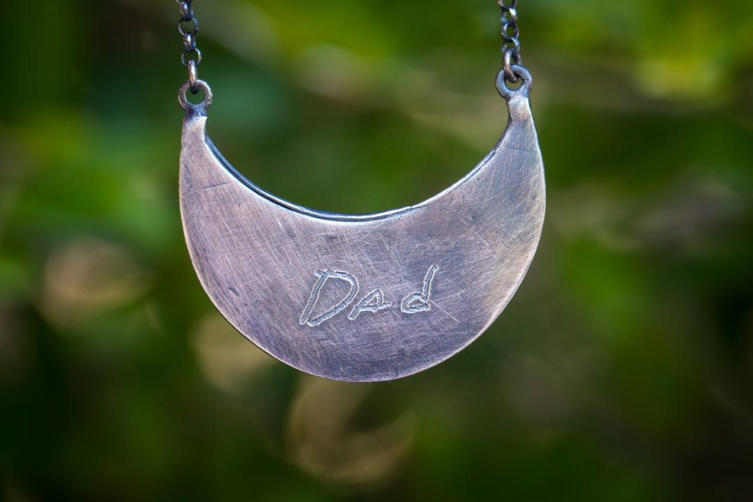 Close-up of the crescent moon pendant with Dad engraved on the brushed silver surface hanging from a dark chain.