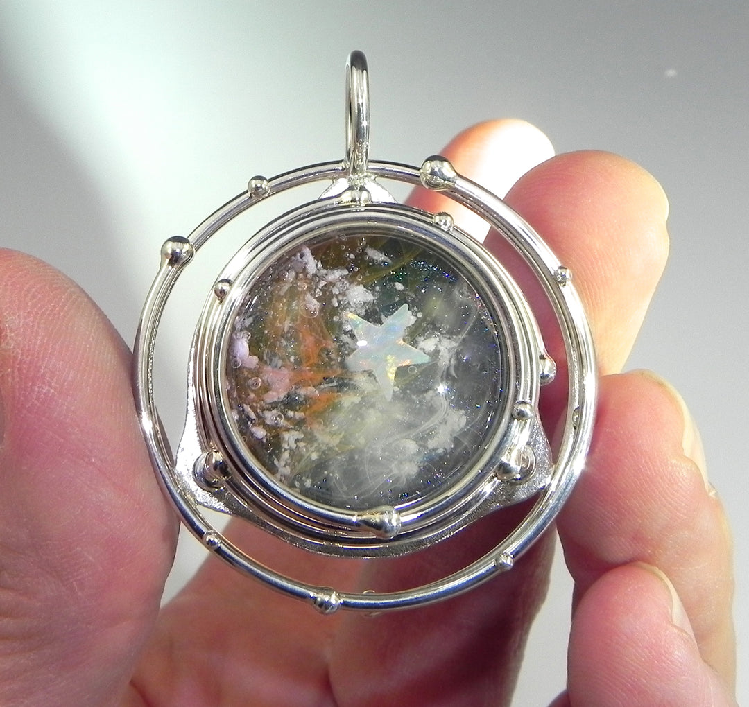 Close up of a silver circular caged pendant with a glass galaxy cabochon and an opal inside, held between fingers.