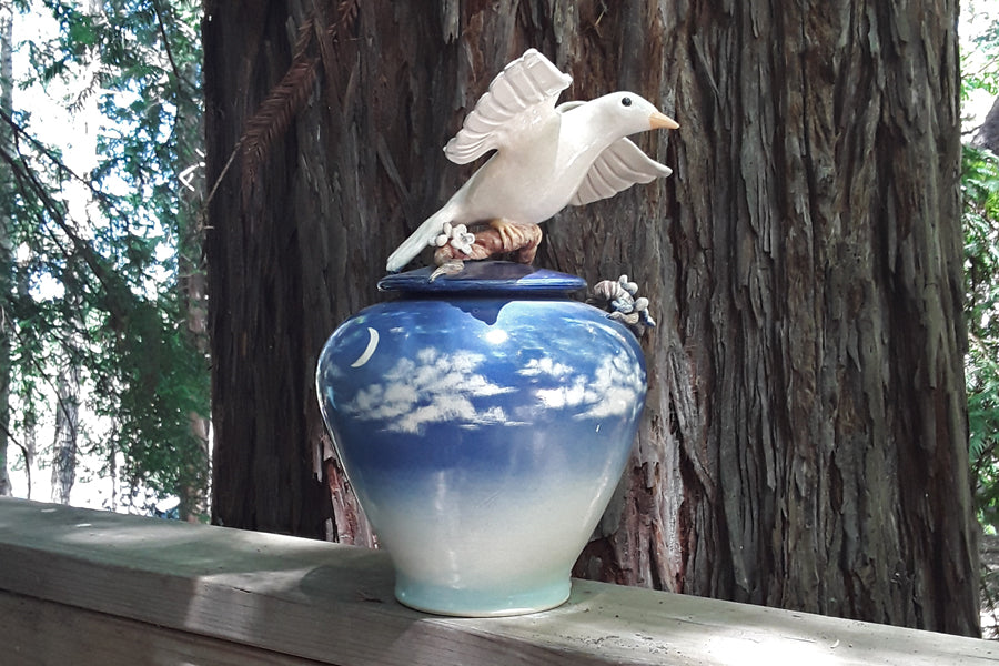 Dove ceramic urn with a blue sky gradient body and a dove sculpture atop the lid, wrapped by a curling branch