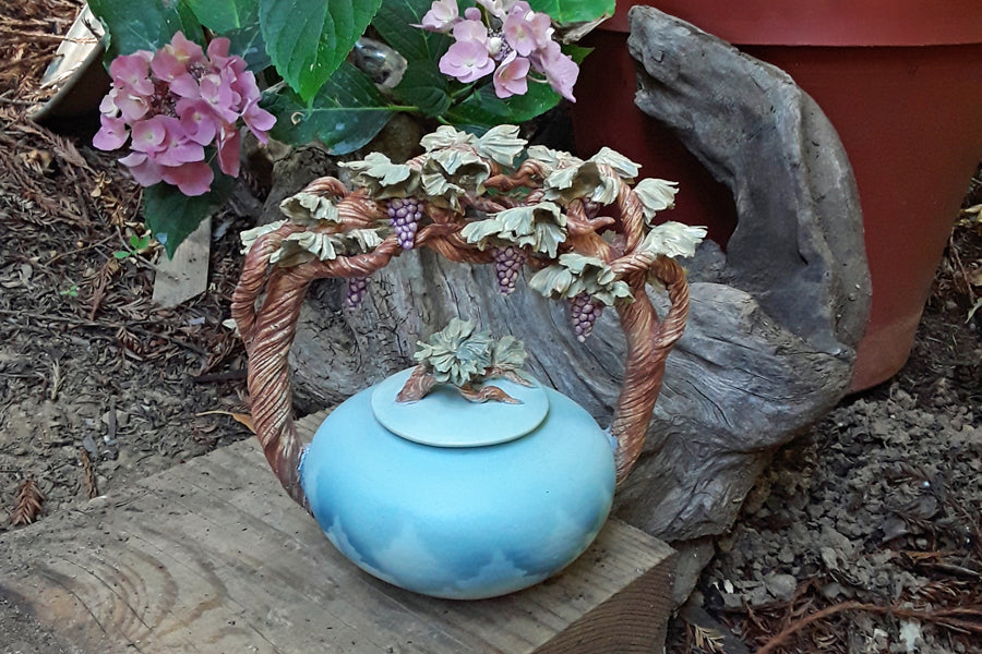 Blue cloud-painted urn with grapevine arch handles and grape clusters, shown against a garden backdrop.