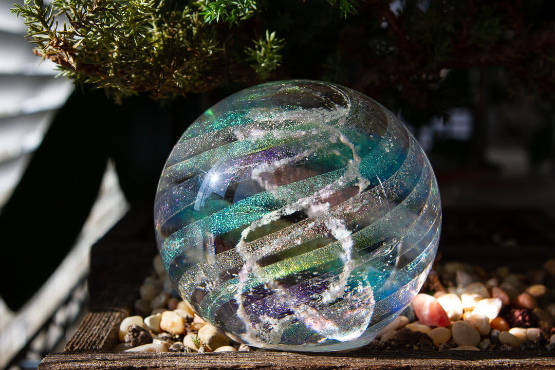 Dichroic rainbow cremation orb outdoors on a wooden ledge with pebbles, iridescent ribbons visible.