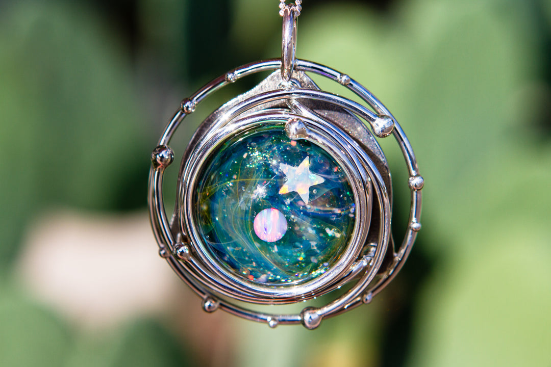 Close-up of a silver orbital pendant featuring a teal galaxy glass cabochon with a star and a planet inside