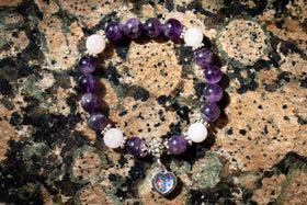 The photo depicts an overhead view of the Amethyst and Rose Quartz Healing Bracelet with Cremation Ash sitting atop a granite surface. The dainty heart charm, made with green and blue opal, faces towards the viewer. Rose quartz bracelet, rose quartz jewelry, amethyst bracelet, amethyst jewelry, natural stone jewelry, remembrance jewelry, jewelry for cremation ash, bracelet for ash