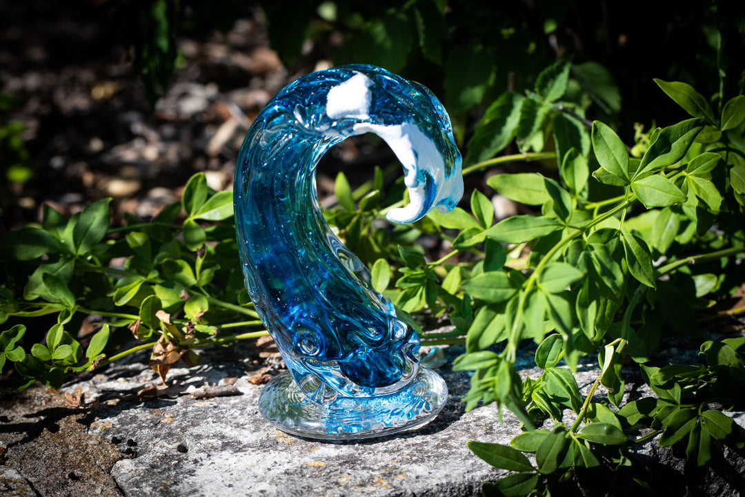 Spirit Wave blue glass paperweight with cremains resting on a stone surface among green plants.