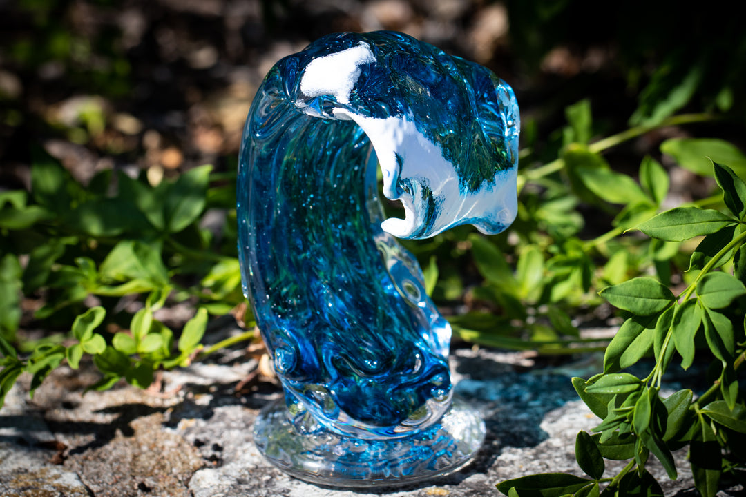Blue Spirit Wave glass sculpture with embedded cremation ashes displayed outdoors on a stone surface among green plants