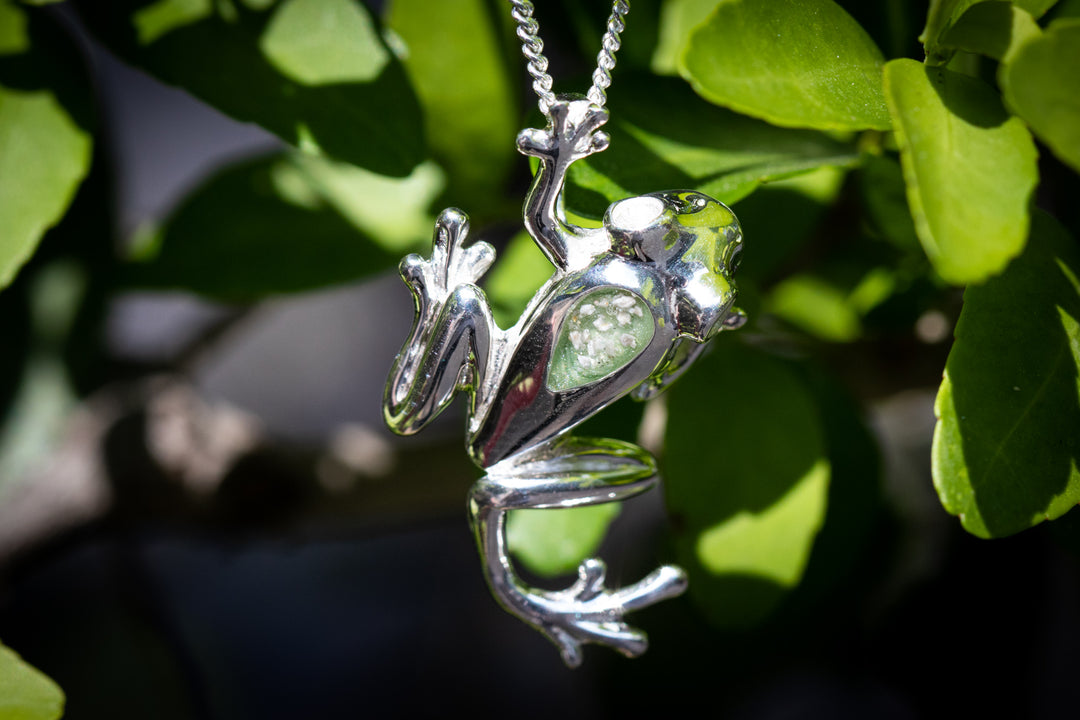Sterling silver Mica Tree Frog Pendant with cremation ash in Pistachio mica shown on a chain against a leafy backdrop.