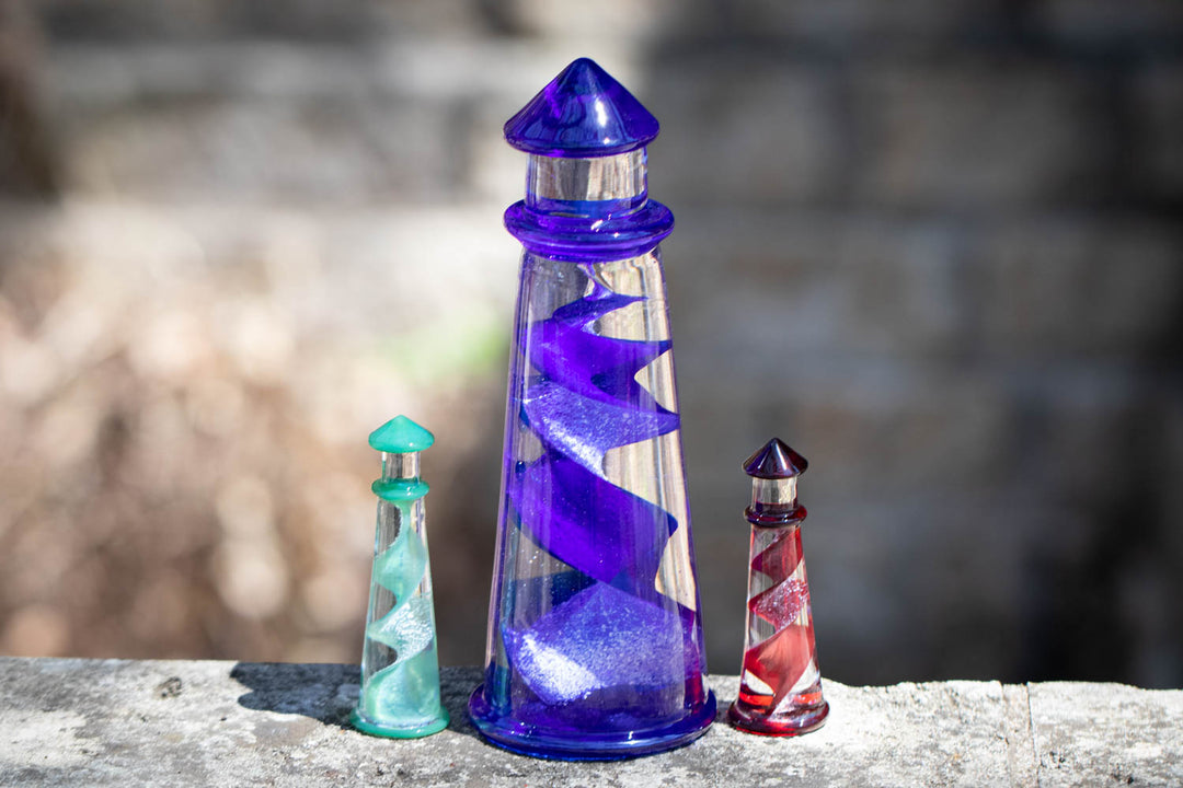 Three colorful memorial glass lighthouses displayed outdoors with a tall purple centerpiece and turquoise and red accents.