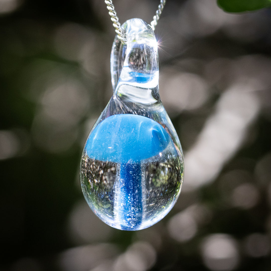 Clear teardrop glass pendant containing a blue mushroom and cremation ash, suspended from an adjustable silver cord