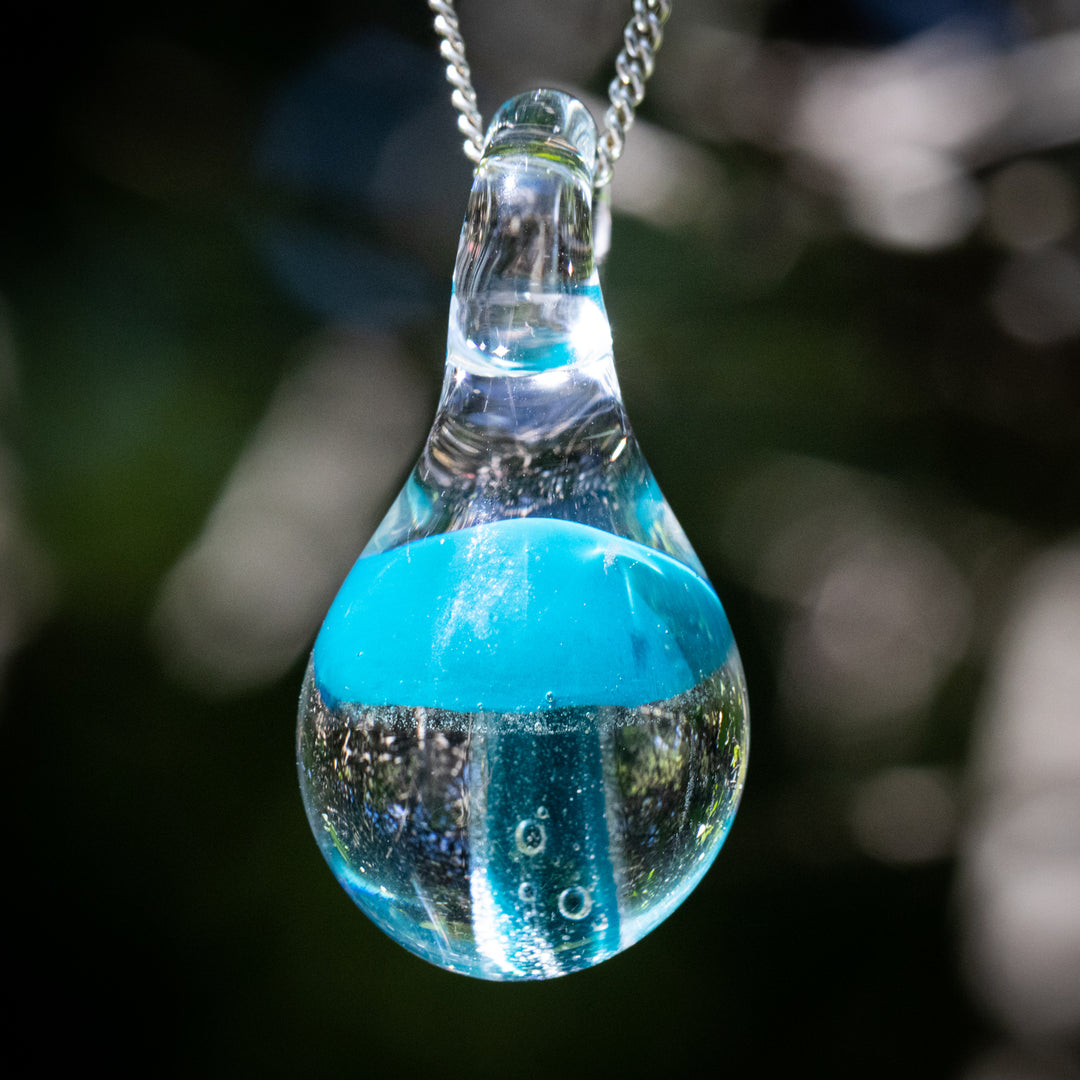Closeup of a teardrop glass pendant showing a blue mushroom inside, infused with cremation ash and hanging from a silver chain