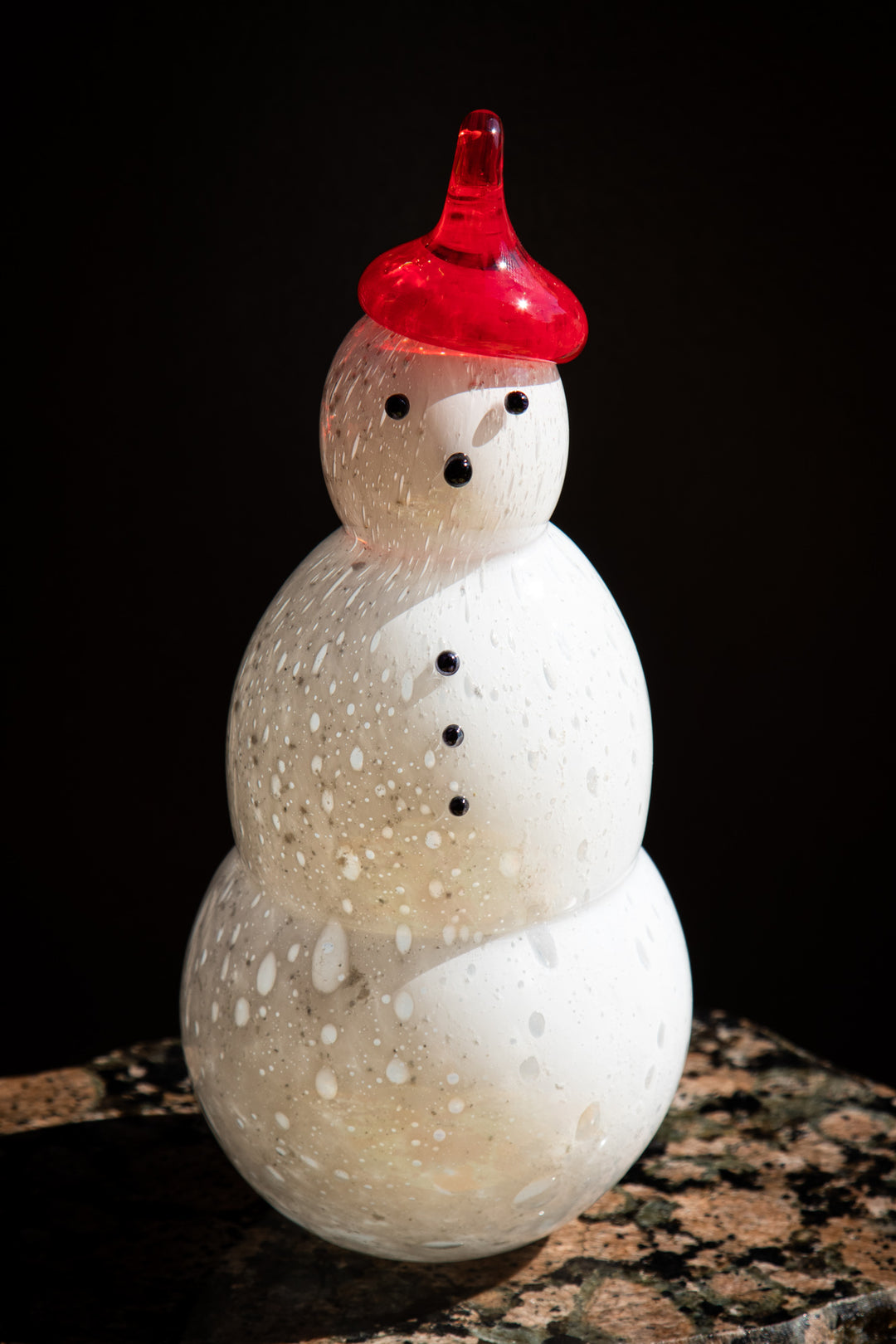 Single white friendly memorial snowman with a red hat featuring cremation ash glaze placed on a textured rock surface.