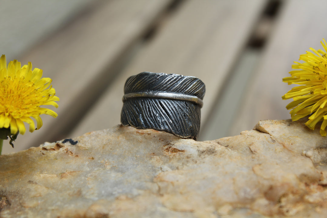 Silver feather ring with a textured feather surface rests on a rock between bright yellow flowers