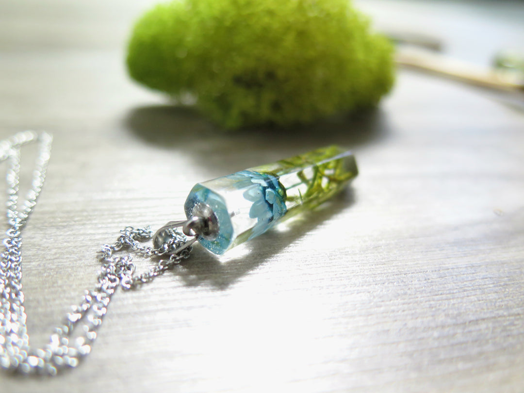 Crystal resin pendant featuring blue starflower and moss resting on a wooden surface with a silver chain nearby