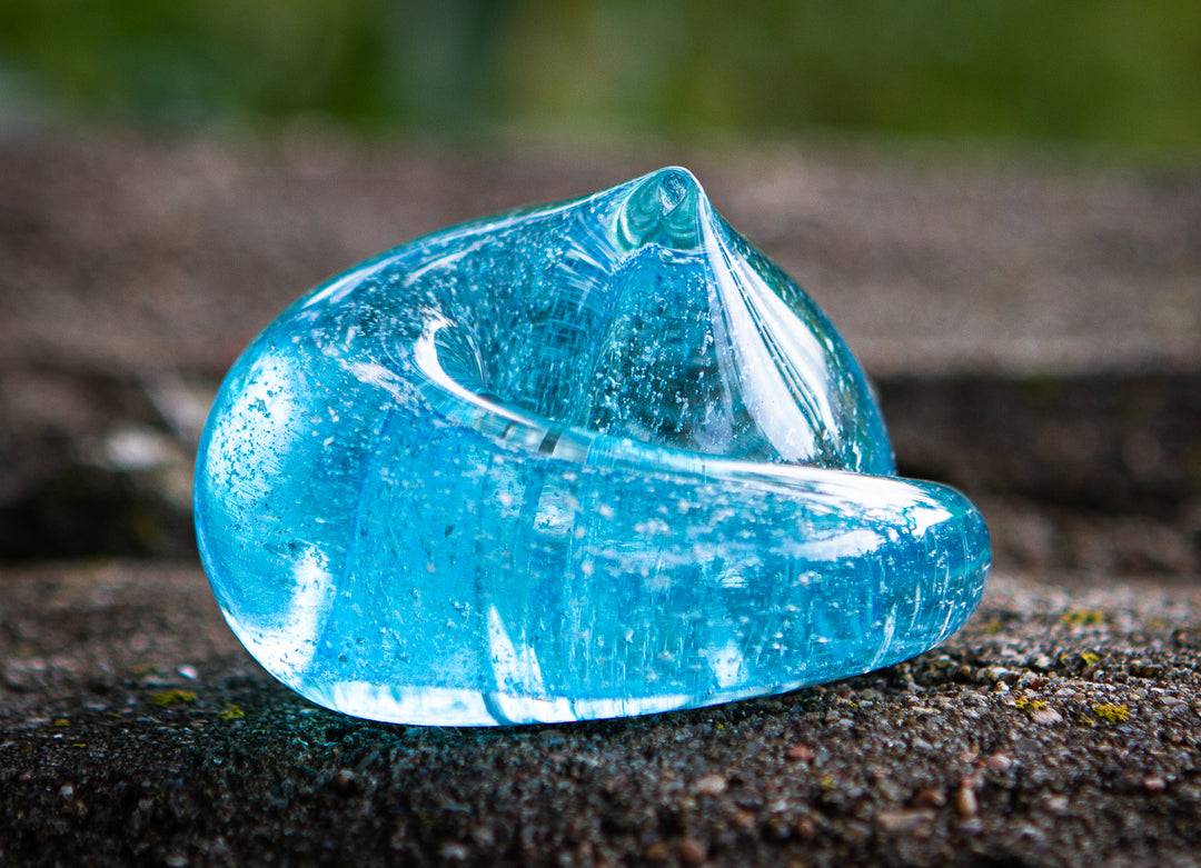 Blue abstract glass sculpture with a curling loop resting on a rough outdoor surface