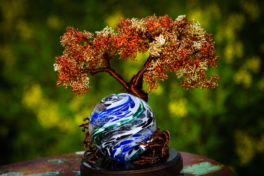 Hand-twisted copper wire tree with a swirling blue and white glass orb at its base on a wooden stand.